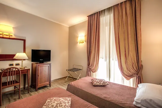 A hotel bedroom with twin beds, a wooden desk and chair, a television on a wooden cabinet, and a luggage stand beside tall curtained windows.