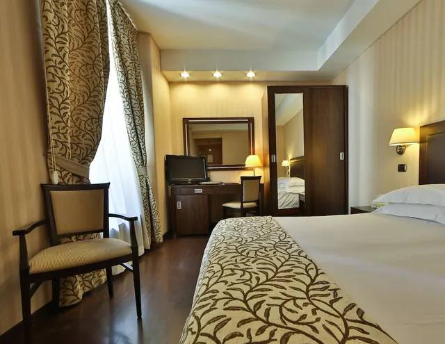 A hotel bedroom with a large bed, an armchair by the window, and a desk with a television and mirror.