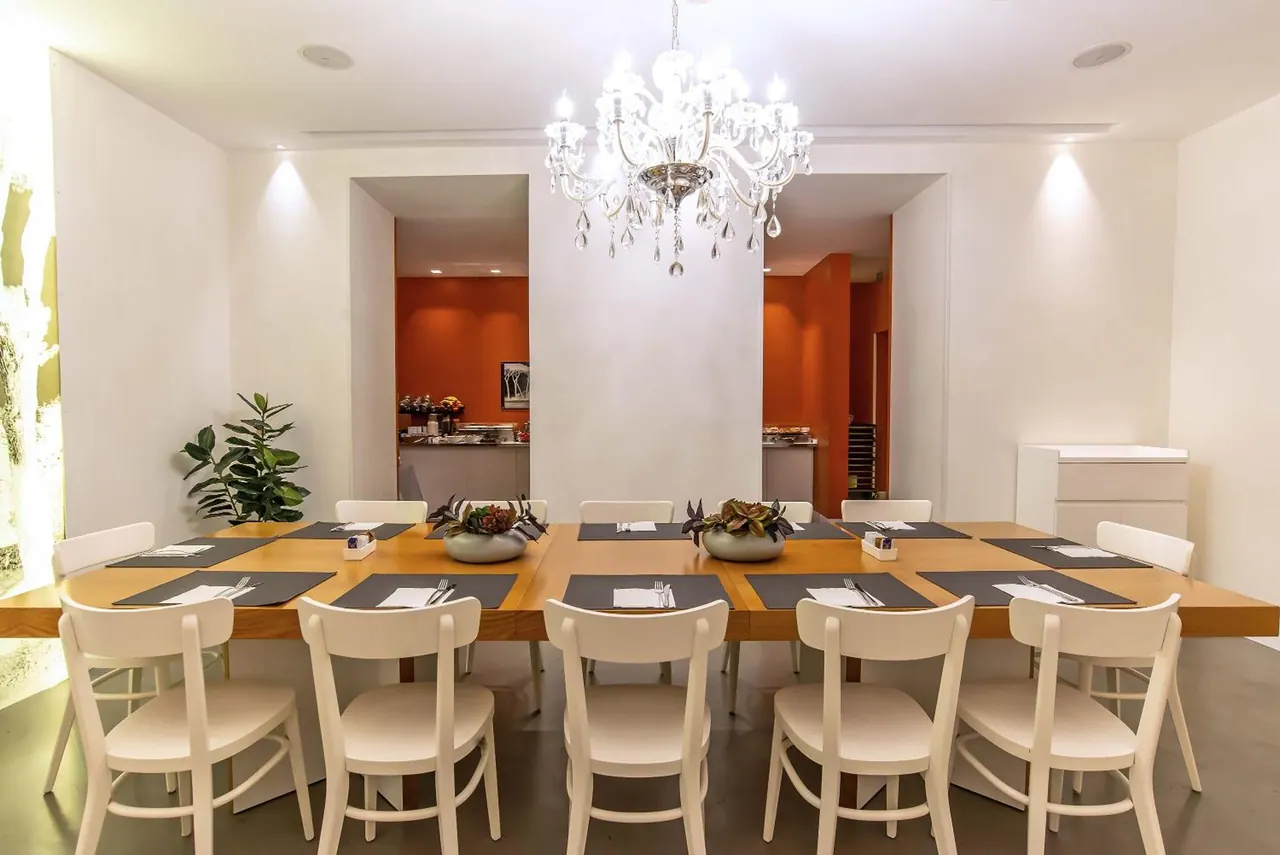A dining room with a long wooden table set with dark placemats and cream chairs, a crystal chandelier overhead, and an orange-walled buffet area in the background.