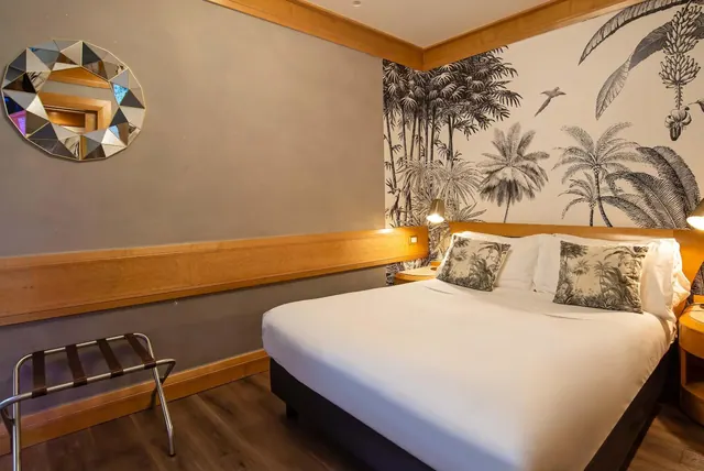 A double bed with white linen and pillows sits beside a wall covered in black and white palm tree wallpaper, with a luggage stand and a faceted round mirror on the grey wall.
