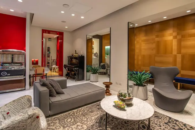 A lounge area with a grey sofa, a grey armchair, a marble coffee table, a jukebox, and an upright piano beside a red wall.