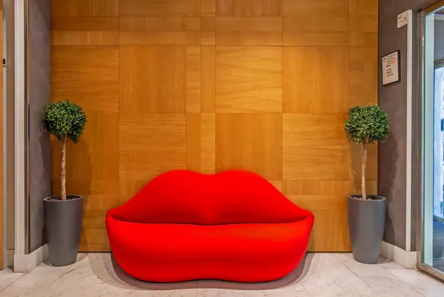 A red lip-shaped sofa stands against a wood-panelled wall between two potted topiary trees in a lobby with a marble floor.
