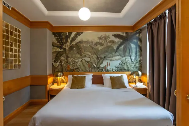 A bedroom with a made-up king bed, green cushions, bedside lamps, and a tropical foliage mural on the wall behind the headboard.