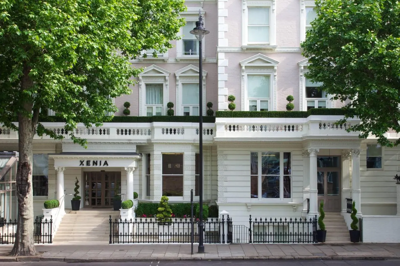 The white stucco front of the Xenia hotel building, with a sign above the entrance, black railings, steps and a street lamp on a tree-lined street.