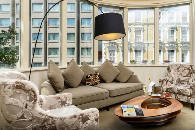 A living area with a beige sofa, patterned armchairs, a black arc floor lamp and a round wooden coffee table, with tall windows looking onto neighbouring buildings.