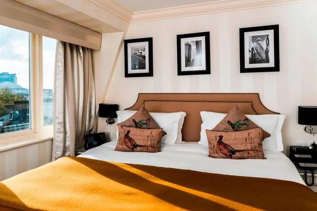 A made-up double bed with decorative cushions and an orange throw sits beside a window that looks out over city buildings, with framed black-and-white city prints on the wall above.