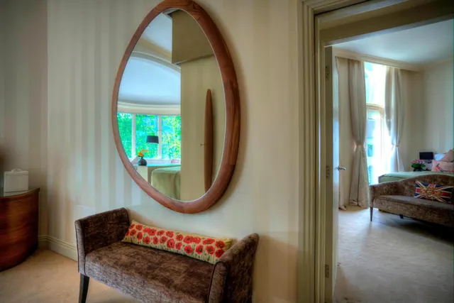 An oval wooden-framed mirror hangs above an upholstered bench with a poppy-print cushion, beside an open door showing a bedroom with a sofa, a bed and a curtained window.