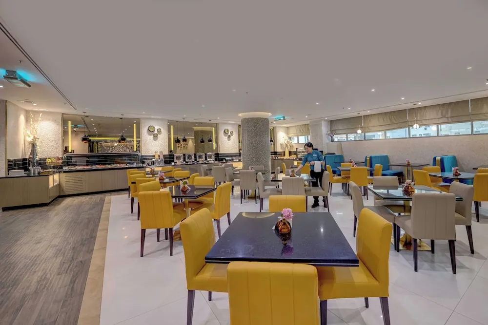 A dining room with yellow chairs and dark square tables, a buffet counter on the left, and a staff member in a blue uniform walking between tables.