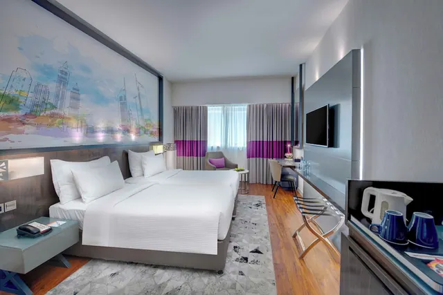 A hotel bedroom with two beds dressed in white linen, a large skyline mural on the wall, a desk, an armchair, and a wall-mounted television.