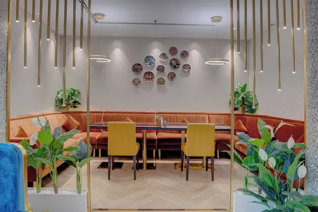 A restaurant seating area with a curved orange velvet banquette, yellow chairs at a dining table, decorative plates on the wall and gold pendant lights above.