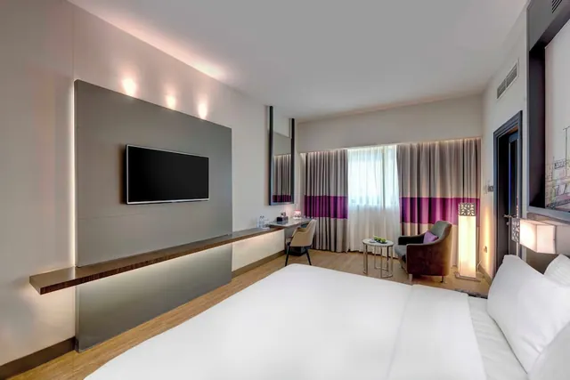 A hotel bedroom with a large white bed, a wall-mounted flat-screen television, a desk with a chair, and an armchair beside a window with purple and grey curtains.