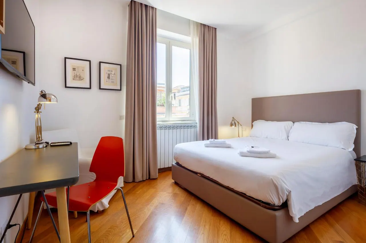 A bedroom with a double bed, a wall-mounted television, a desk with a lamp and a red chair, and a window looking onto neighbouring buildings.