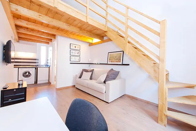 A living area with a white sofa under framed pictures, a wall-mounted television, a wooden staircase to a loft and a small kitchenette with a washing machine in the background.