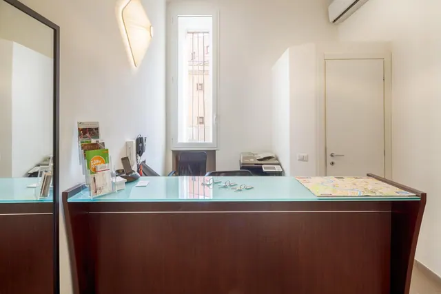 A wooden reception desk with a glass top holds leaflets, keys and a folded map, with a barred window, printer and wall air conditioner in the small room behind it.