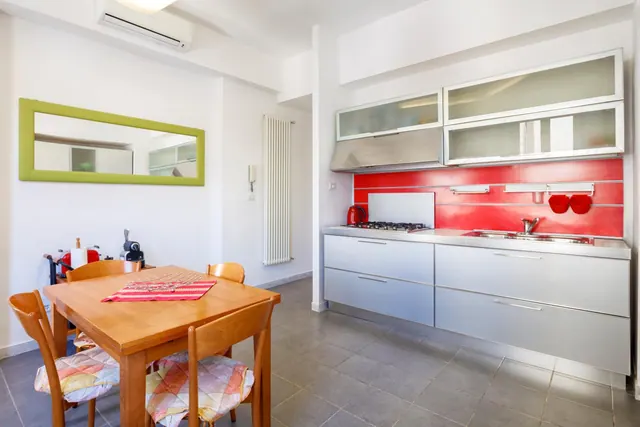 A kitchen corner with a red backsplash, a gas hob and sink beside white cabinets, and a wooden dining table with four chairs.