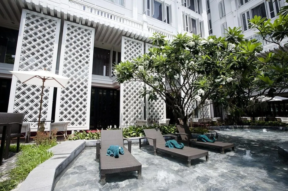 Two wicker loungers with folded teal towels stand in shallow pool water beside a white lattice-screened building and a frangipani tree, with a white umbrella at left.