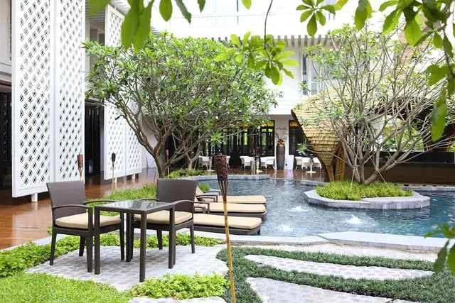An outdoor pool area with sun loungers, a wicker table and chairs, frangipani trees and a thatched pavilion beside the water.