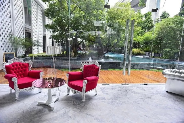 Two red tufted armchairs and a round side table stand on a marble floor in front of a glass wall overlooking a pool and trees.