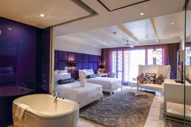 A hotel bedroom with two white beds, a freestanding bathtub in the foreground, a wingback sofa, and a purple glass partition wall.