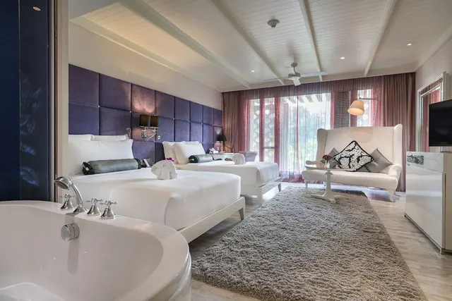 A bedroom with two beds, a freestanding bathtub in the foreground, a white wingback chair, a shag rug and sheer curtains over floor-to-ceiling windows that look out onto greenery.
