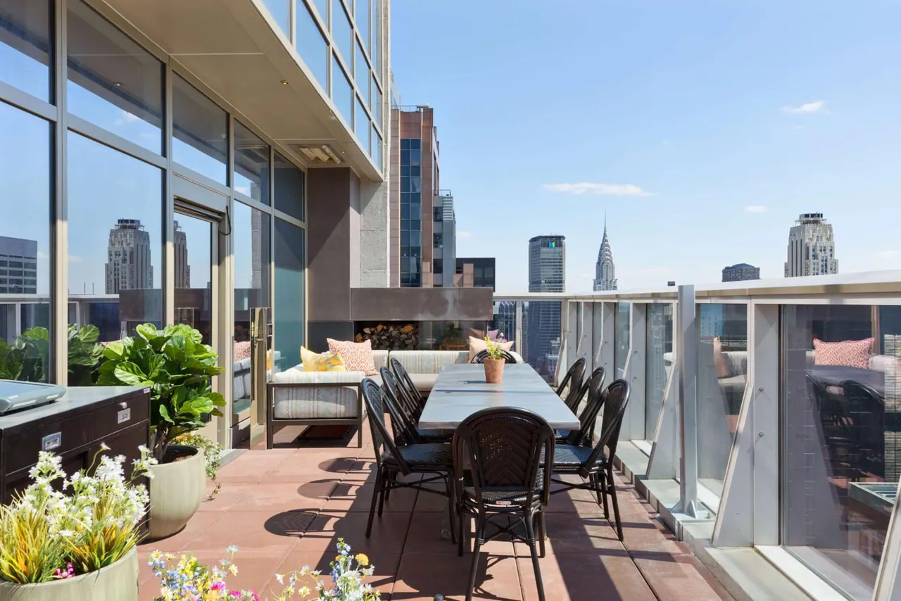 A rooftop terrace with a long dining table, black chairs, lounge seating and planters, with city towers and the Chrysler Building visible beyond the glass railing.