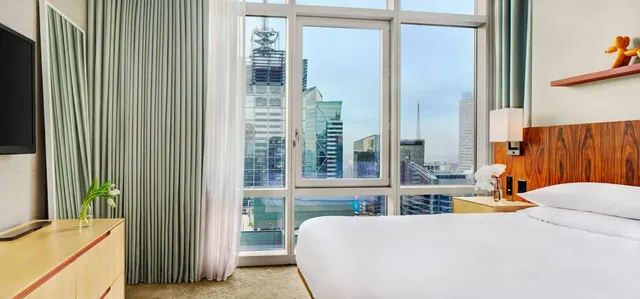 A king bed with white linen sits in a hotel room beside floor-to-ceiling windows overlooking a city of tall buildings.