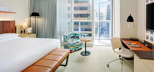 A bedroom with a king bed, a patterned armchair, a desk with a chair, and an open glass door onto a balcony with high-rise buildings beyond.