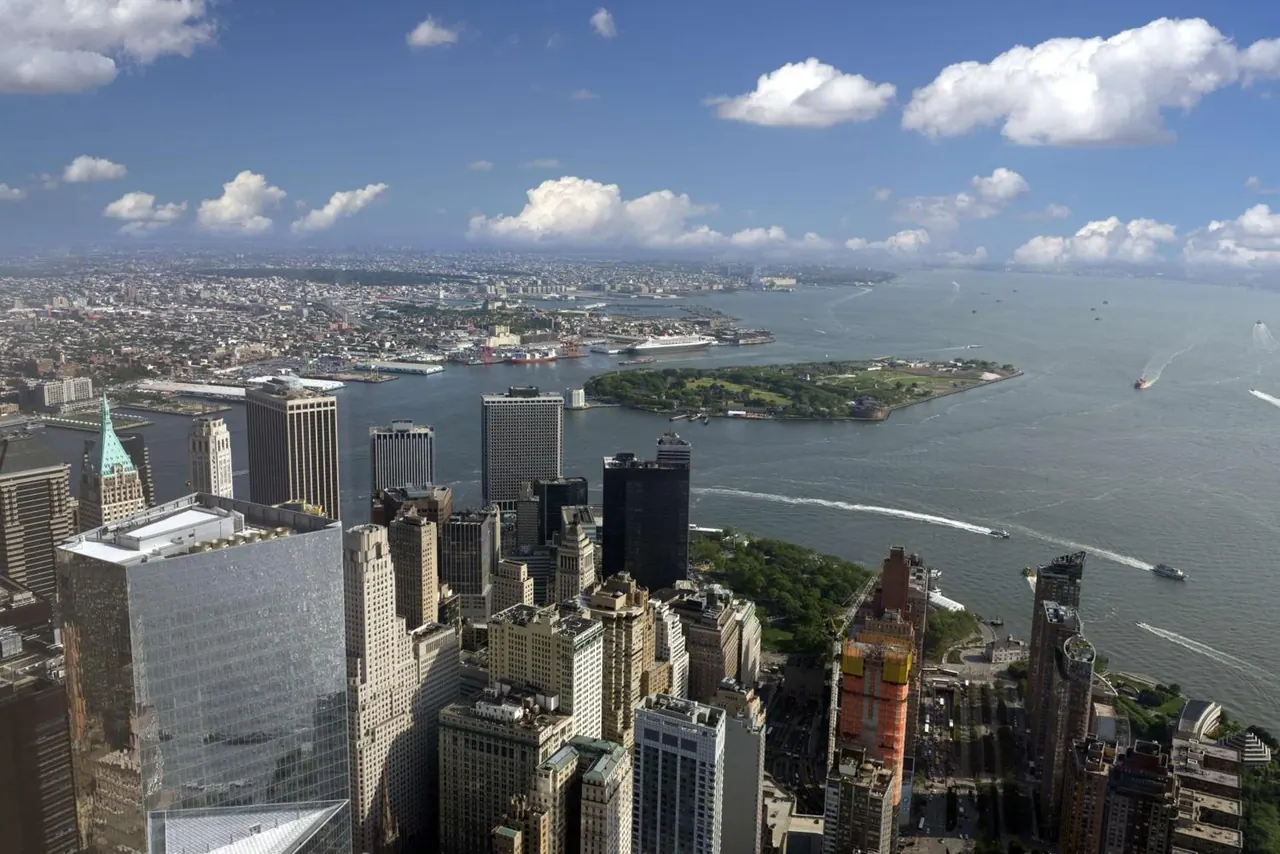 Aerial view over lower Manhattan skyscrapers, New York Harbor, and Governors Island under a partly cloudy sky.