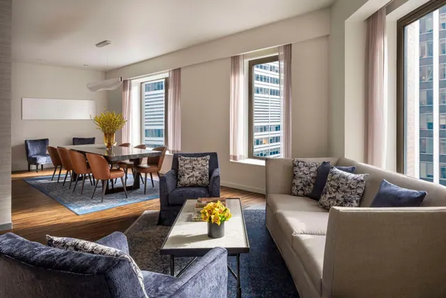 A staged living area with a beige sofa, navy armchairs and a coffee table with yellow flowers, with a dining table beyond and windows overlooking neighbouring high-rise buildings.
