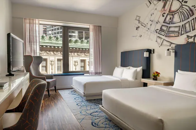 A hotel room with two beds, a wall-mounted television, a desk, and an armchair by a window looking out at an ornate stone building.
