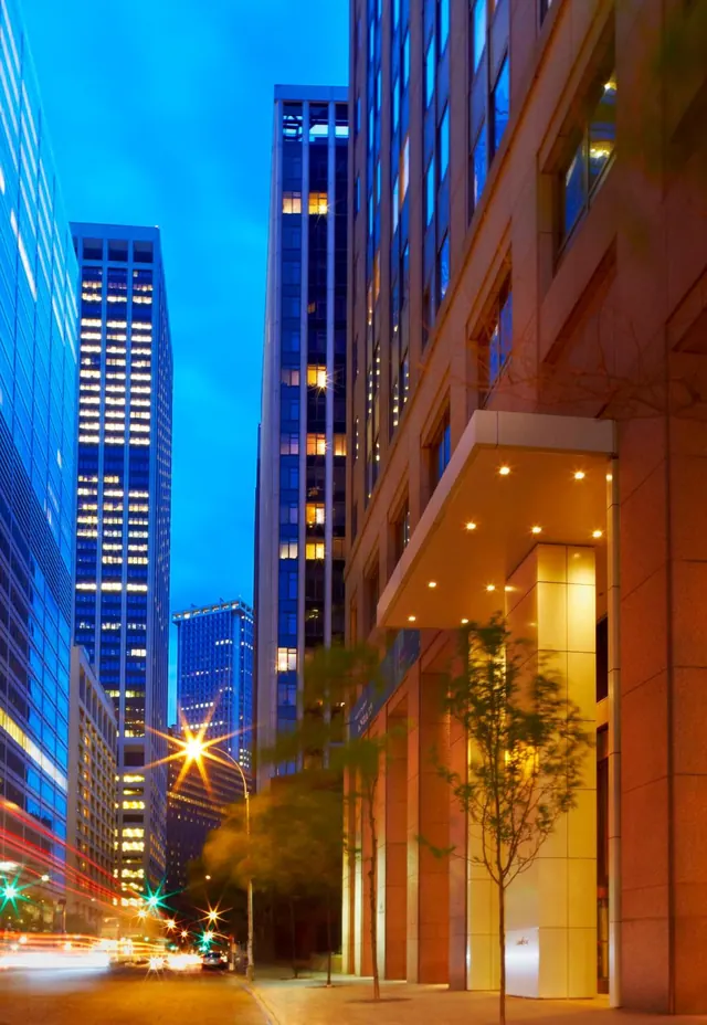 Dusk view of a city street between tall glass office towers, with a warmly lit building entrance canopy and young trees on the sidewalk.