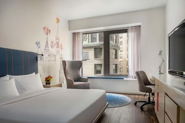 A hotel bedroom with a king bed, a grey wingback armchair, a corner desk with a chair, a wall-mounted television, and a window looking onto neighbouring brick buildings.