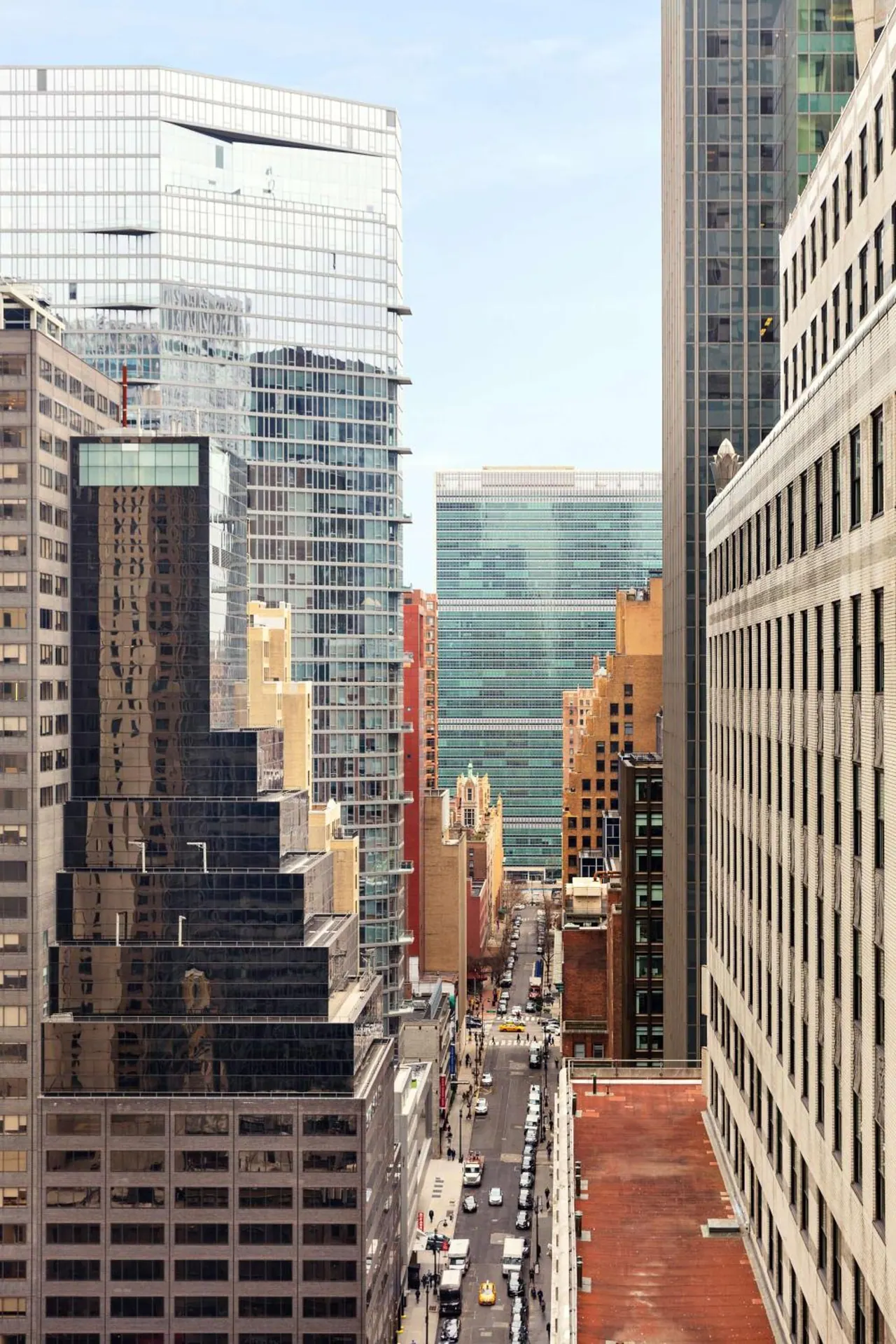 A view down a narrow city street between tall glass and stone office towers, with traffic and a yellow taxi visible far below.