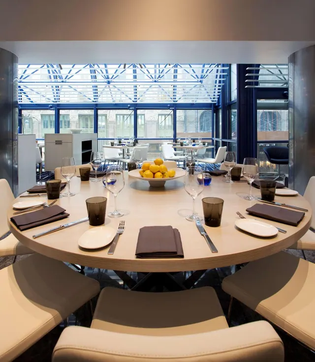 A round dining table is set with plates, glasses, napkins and a bowl of lemons in a restaurant with floor-to-ceiling windows and a glass roof.