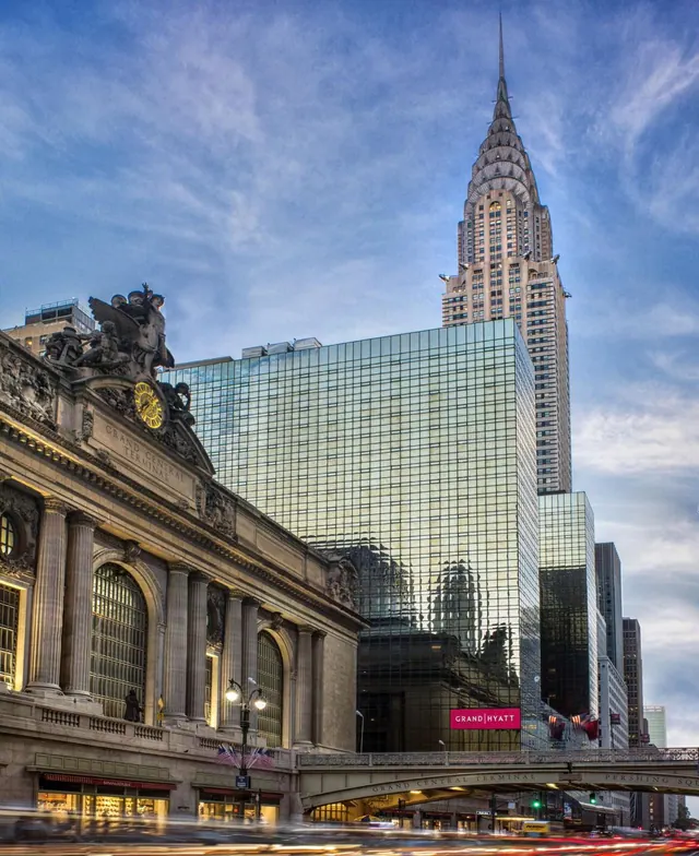 Exterior view of the Grand Hyatt glass tower beside Grand Central Terminal, with the Chrysler Building spire rising behind it at dusk.