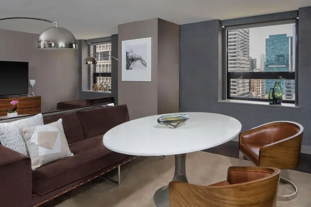 A living area with a brown velvet sofa, a white oval table with leather chairs, a flat-screen television, and a window overlooking city skyscrapers.