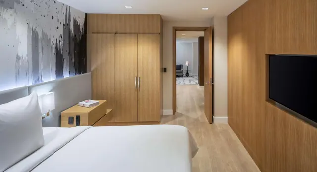 A bedroom with a made white bed, a wood wardrobe, a wall-mounted television and an open door to an adjoining sitting area.
