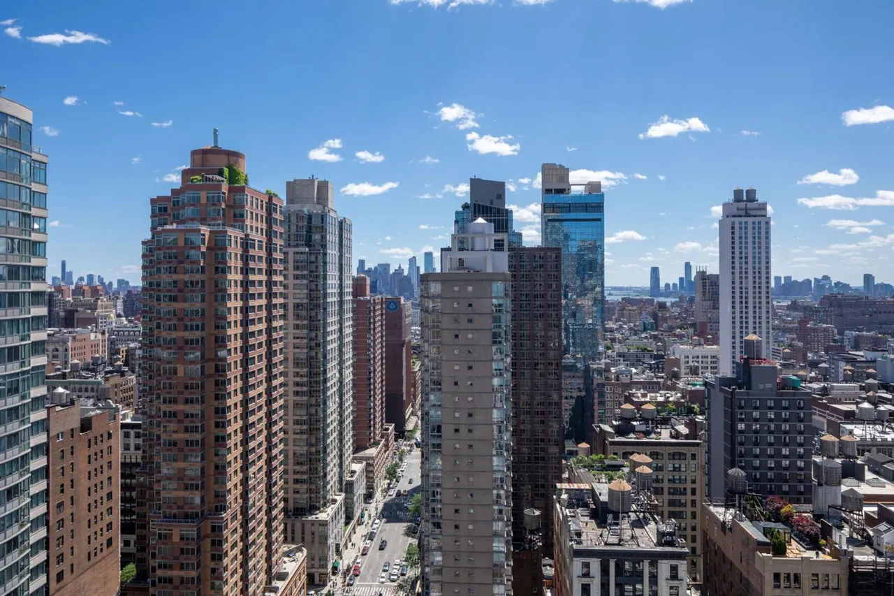 Aerial view over a dense cluster of high-rise towers and rooftops under a blue sky with scattered clouds.
