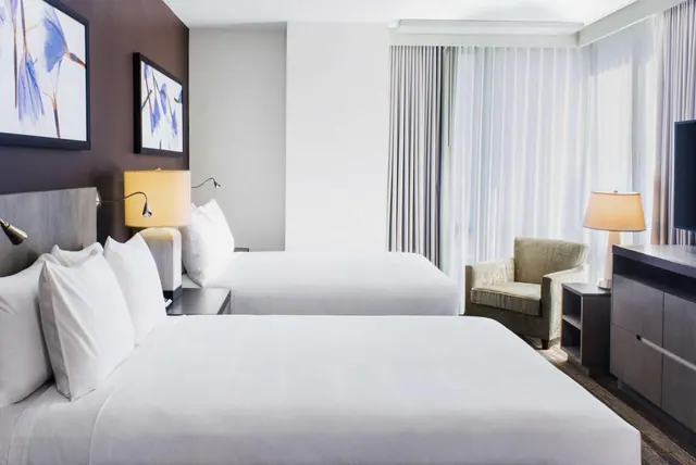 A hotel bedroom with two made beds with white bedding, an armchair, bedside lamps, a dresser and a wall-mounted television, with sheer curtains behind.