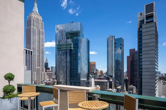 A rooftop terrace with wooden tables and chairs overlooks Midtown Manhattan skyscrapers, including the Empire State Building.