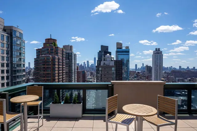 A rooftop terrace with wooden tables and chairs and white planters overlooks a city skyline under a clear blue sky.