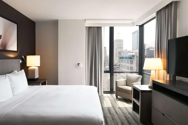 A king bed with white linen, two bedside lamps, an armchair and a wall-mounted television face floor-to-ceiling windows that look out over city buildings.