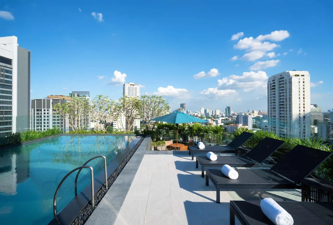 A rooftop swimming pool with sun loungers, white towels and a teal umbrella, with high-rise buildings of the city beyond.
