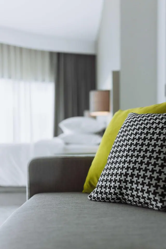 A grey sofa with a lime green cushion and a black and white houndstooth cushion sits in the foreground, with a made bed and a lamp blurred in the background.
