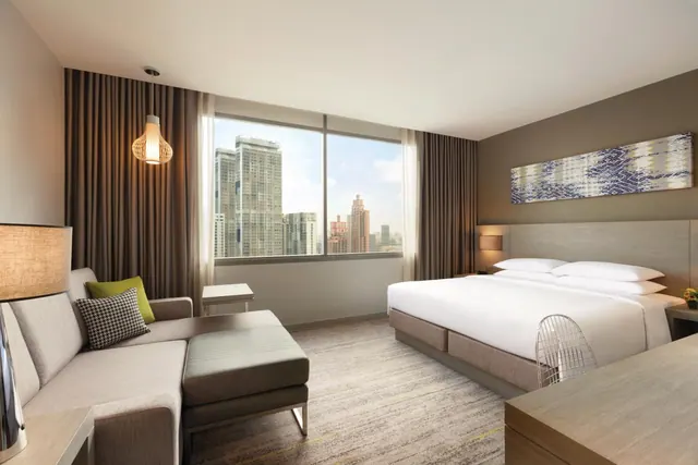 A bedroom with a king bed, a grey sofa and ottoman, and a large window looking out over city high-rise buildings.