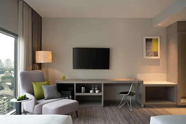 A hotel living area with a grey armchair and ottoman, a wall-mounted television, a console desk with a kettle and cups, and a window showing city buildings.