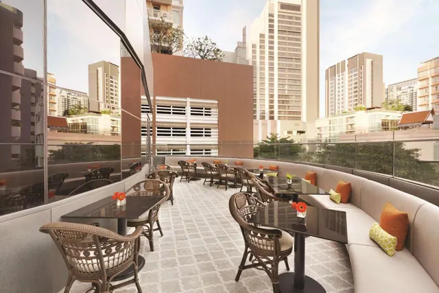 An outdoor terrace with wicker chairs, dark tables and a curved cushioned bench overlooks high-rise buildings under a pale sky.