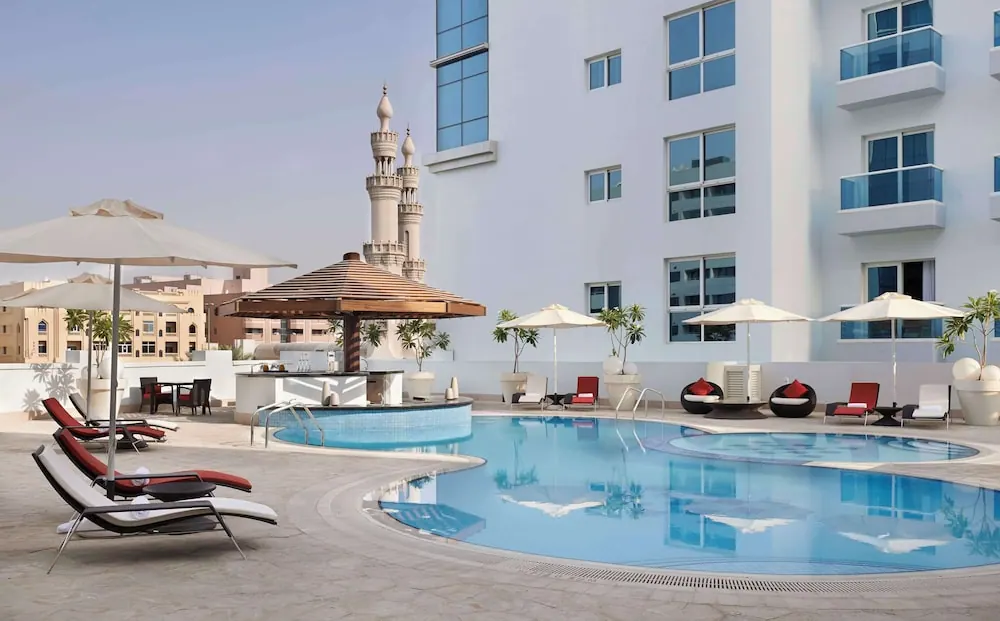 An outdoor pool with loungers and umbrellas on a stone deck beside a white hotel building, with a minaret visible in the background.