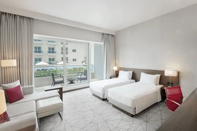 A hotel room with two white single beds, a grey sofa and a sliding glass door opening onto a balcony with chairs and a view of a neighbouring building.