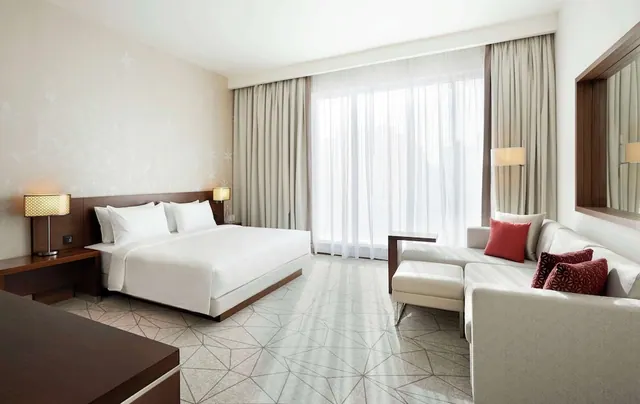 A hotel bedroom with a king bed, a grey sofa with red cushions, and a wall of sheer-curtained floor-to-ceiling windows.